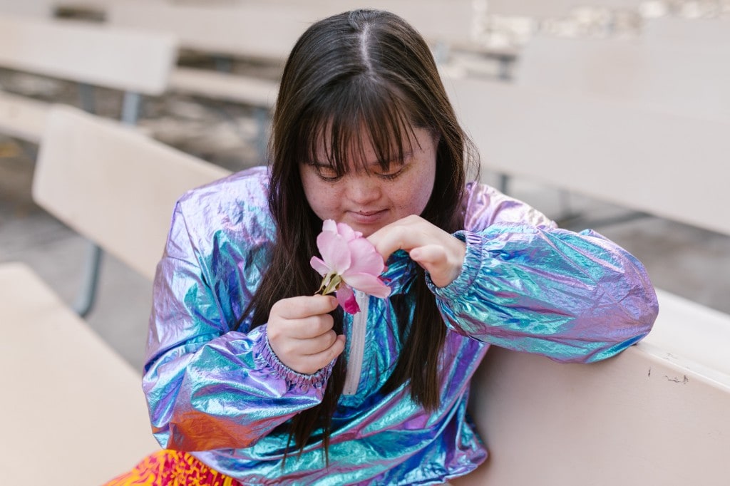 A young woman with Down syndrome in an iridescent jacket, gently holding a pink rose while seated on a bench outdoors