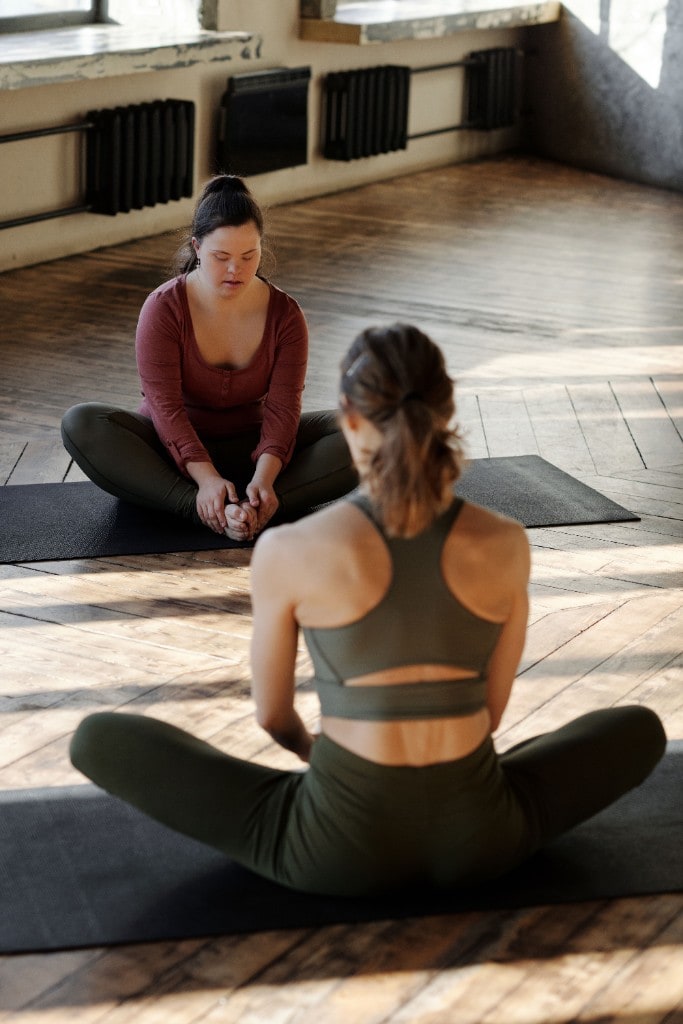 Two women practicing yoga together in a butterfly stretch on mats in a sunlit studio